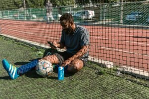 Athlete taking a break on a soccer field with ball and smartphone, enjoying leisure time.